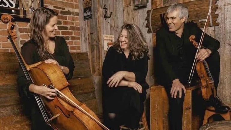 Trois musiciens vêtus de noir discutent et sourient dans un décor rustique aux murs de bois et de briques. Une femme tient un violoncelle, tandis qu’un homme tient un violon. Une autre femme est assise entre eux. L’ambiance est chaleureuse et conviviale