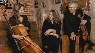 Trois musiciens vêtus de noir discutent et sourient dans un décor rustique aux murs de bois et de briques. Une femme tient un violoncelle, tandis qu’un homme tient un violon. Une autre femme est assise entre eux. L’ambiance est chaleureuse et conviviale