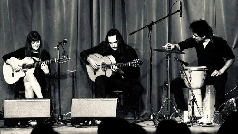 Photo en noir et blanc de trois musiciens sur scène devant un rideau. Deux personnes jouent de la guitare acoustique tandis qu’une troisième joue des percussions. Les artistes, vêtus de noir, sont assis sous des microphones dans une ambiance intimiste rappelant un spectacle de flamenco.