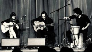 Photo en noir et blanc de trois musiciens sur scène devant un rideau. Deux personnes jouent de la guitare acoustique tandis qu’une troisième joue des percussions. Les artistes, vêtus de noir, sont assis sous des microphones dans une ambiance intimiste rappelant un spectacle de flamenco.