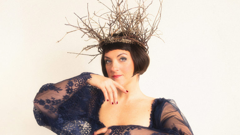 Portrait artistique d’une femme portant une robe bleu foncé aux manches transparentes en dentelle. Elle pose devant un fond clair avec une couronne faite de branches entrelacées sur la tête. Son maquillage et sa posture donnent une ambiance élégante et théâtrale.