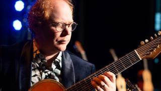 Un musicien joue d’une guitare acoustique à plusieurs cordes sur une scène éclairée de lumière bleue et chaude. Il porte des lunettes rondes, un veston foncé et une chemise à motifs. D’autres guitares apparaissent floues en arrière-plan, créant une ambiance de concert intimiste.