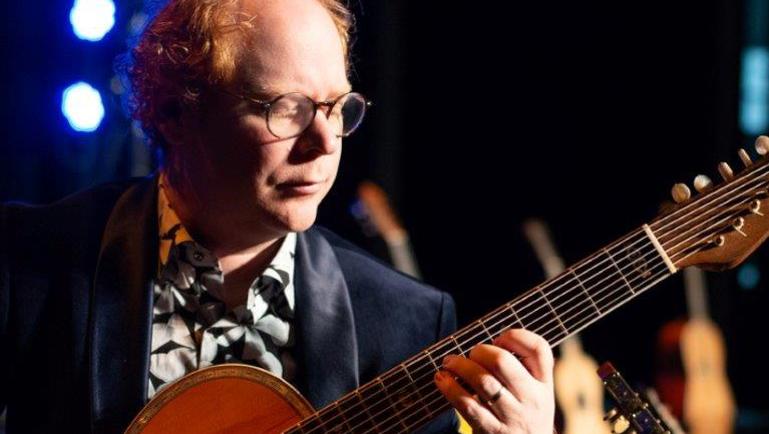 Un musicien joue d’une guitare acoustique à plusieurs cordes sur une scène éclairée de lumière bleue et chaude. Il porte des lunettes rondes, un veston foncé et une chemise à motifs. D’autres guitares apparaissent floues en arrière-plan, créant une ambiance de concert intimiste.