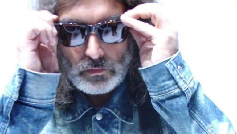 Portrait rapproché d’un homme portant des lunettes de soleil et une veste en denim bleu pâle. Il ajuste ses lunettes avec ses deux mains et porte une guitare électrique en bandoulière. Le fond blanc très lumineux donne à l’image un style épuré et contrasté.