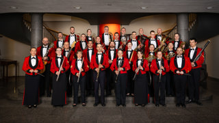 Photo de groupe d’un ensemble musical militaire en uniforme rouge et noir, posant à l’intérieur d’un bâtiment. Les musiciens, disposés sur plusieurs rangées, tiennent divers instruments à vent et percussions, dont des clarinettes, flûtes, trombones, tubas, cors français et un basson. L’arrière-plan présente un escalier symétrique et des colonnes sombres, avec un éclairage chaleureux au centre