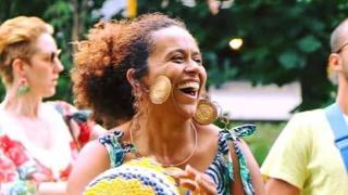 Une femme souriante joue d’un instrument de percussion tressé jaune et bleu lors d’un événement extérieur. Elle porte une robe à motifs colorés et de grandes boucles d’oreilles rondes. Des personnes floues et de la végétation apparaissent en arrière-plan, créant une ambiance festive et conviviale.