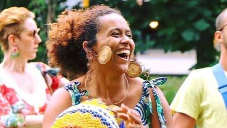 Une femme souriante joue d’un instrument de percussion tressé jaune et bleu lors d’un événement extérieur. Elle porte une robe à motifs colorés et de grandes boucles d’oreilles rondes. Des personnes floues et de la végétation apparaissent en arrière-plan, créant une ambiance festive et conviviale.