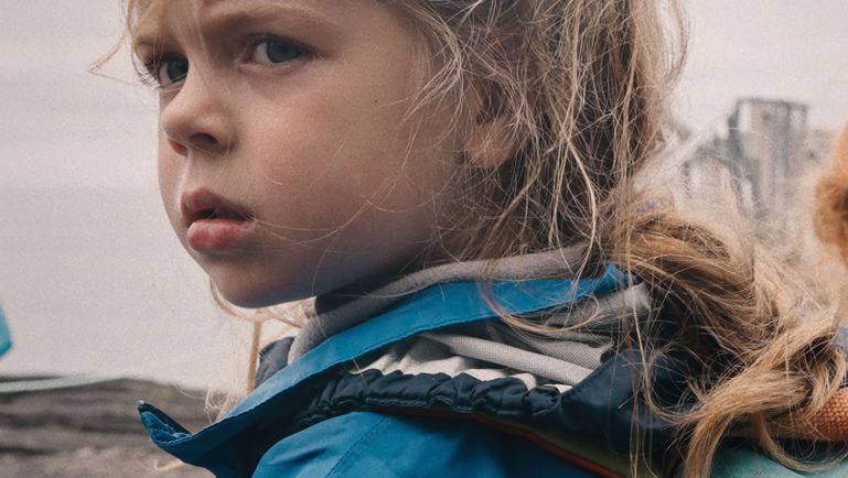 Affiche de film montrant une jeune fille blonde portant un manteau bleu et un sac à dos, regardant vers l’arrière avec une expression sérieuse. Le titre “À hauteur d’enfant” apparaît en écriture blanche manuscrite au-dessus d’elle. Le fond est flou et grisâtre, créant une ambiance douce et introspective.