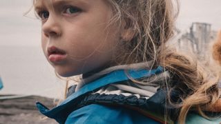 Affiche de film montrant une jeune fille blonde portant un manteau bleu et un sac à dos, regardant vers l’arrière avec une expression sérieuse. Le titre “À hauteur d’enfant” apparaît en écriture blanche manuscrite au-dessus d’elle. Le fond est flou et grisâtre, créant une ambiance douce et introspective.
