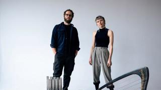 Deux artistes posent debout dans un studio au fond blanc, entourés de plusieurs instruments de musique, dont des handpans, une harpe et un métallophone. L’homme porte des vêtements foncés et des lunettes, tandis que la femme porte un haut noir sans manches et un pantalon gris.
