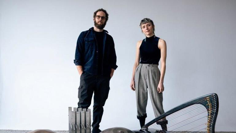 Deux artistes posent debout dans un studio au fond blanc, entourés de plusieurs instruments de musique, dont des handpans, une harpe et un métallophone. L’homme porte des vêtements foncés et des lunettes, tandis que la femme porte un haut noir sans manches et un pantalon gris.