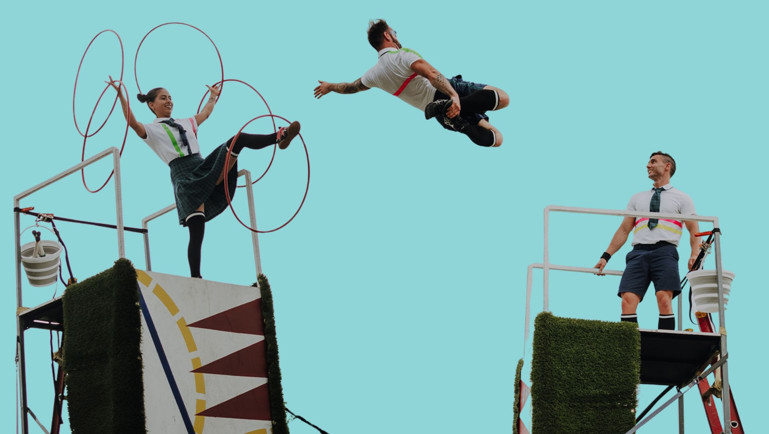Trois artistes de cirque réalisent une performance acrobatique en plein air sur des structures surélevées. Une femme manipule plusieurs cerceaux rouges tandis qu’un homme est suspendu en plein saut entre deux plateformes. Un autre homme observe la scène depuis une structure voisine. Les artistes portent des costumes évoquant des uniformes scolaires, sur fond de ciel bleu clair.