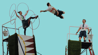 Trois artistes de cirque réalisent une performance acrobatique en plein air sur des structures surélevées. Une femme manipule plusieurs cerceaux rouges tandis qu’un homme est suspendu en plein saut entre deux plateformes. Un autre homme observe la scène depuis une structure voisine. Les artistes portent des costumes évoquant des uniformes scolaires, sur fond de ciel bleu clair.