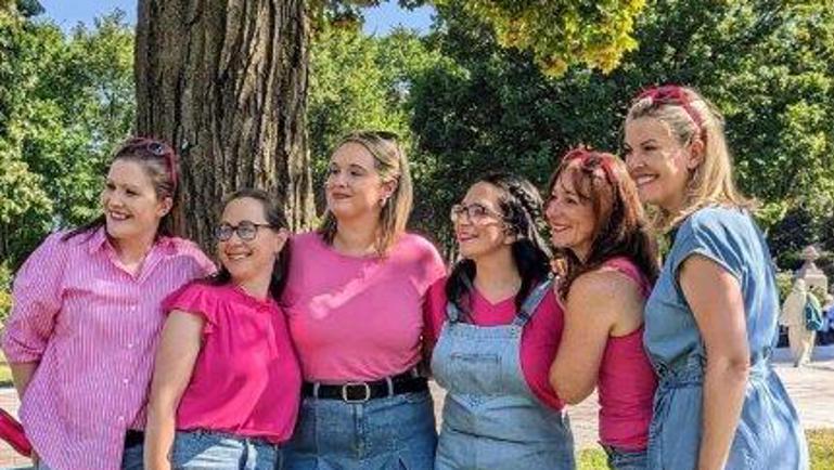 Six femmes posent ensemble dans un parc ensoleillé devant un grand arbre. Elles portent des vêtements dans des tons de rose et de bleu denim, avec des chaussures roses assorties. Elles sourient et regardent dans la même direction, créant une ambiance conviviale et festive.