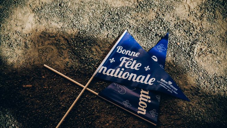 Deux petits drapeaux bleus de la Fête nationale du Québec sont posés sur un sol de gravier et de terre. On peut lire « Bonne Fête nationale » en lettres blanches sur les drapeaux décorés de fleurs de lys. Des traces de pas sont visibles autour.