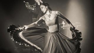 Photo en noir et blanc d’une danseuse en mouvement sur scène, portant une longue robe fluide qu’elle déploie de chaque côté avec ses bras. À l’arrière-plan, un musicien joue de la trompette dans une lumière tamisée. L’ambiance évoque une performance de danse et musique en direct.