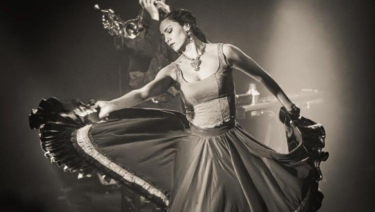 Photo en noir et blanc d’une danseuse en mouvement sur scène, portant une longue robe fluide qu’elle déploie de chaque côté avec ses bras. À l’arrière-plan, un musicien joue de la trompette dans une lumière tamisée. L’ambiance évoque une performance de danse et musique en direct.