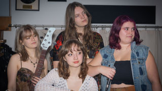 Quatre jeunes musiciennes posent ensemble dans un local de répétition ou un studio. L’une tient une guitare basse blanche, tandis qu’une autre tient un microphone. Les membres du groupe portent des vêtements décontractés et regardent dans différentes directions, créant une ambiance spontanée et artistique