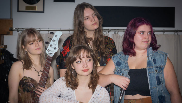 Quatre jeunes musiciennes posent ensemble dans un local de répétition ou un studio. L’une tient une guitare basse blanche, tandis qu’une autre tient un microphone. Les membres du groupe portent des vêtements décontractés et regardent dans différentes directions, créant une ambiance spontanée et artistique