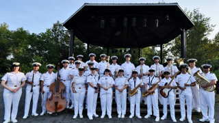 Photo de groupe d’un ensemble musical de la réserve navale posant devant une scène extérieure couverte. Les musiciens portent des uniformes blancs avec casquettes et tiennent divers instruments, dont des clarinettes, saxophones, trombones, trompettes, une contrebasse, une guitare électrique et un tuba. Des arbres et un ciel bleu dégagé apparaissent en arrière-plan.