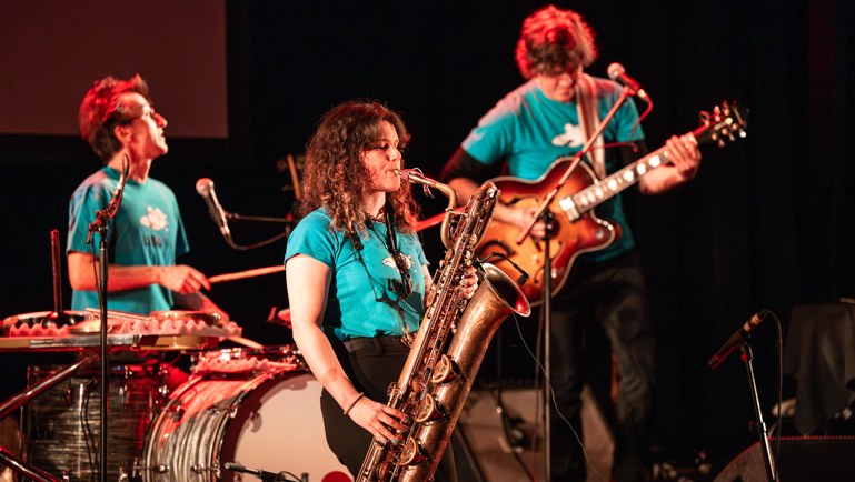Musicien·nes sur scène, avec saxophone baryton, batterie et guitare, lors d’une performance musicale.