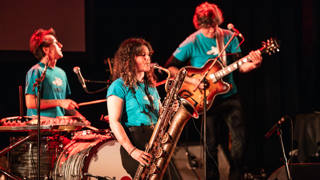 Musicien·nes sur scène, avec saxophone baryton, batterie et guitare, lors d’une performance musicale.