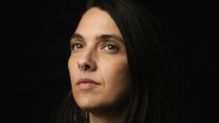Portrait sobre d’une femme aux longs cheveux foncés posé devant un fond noir. Elle regarde l’objectif avec une expression calme et sérieuse. Un anneau au nez et un tatouage partiellement visible au haut du torse ajoutent une touche distinctive à l’image