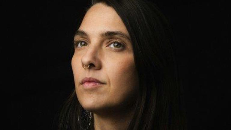 Portrait sobre d’une femme aux longs cheveux foncés posé devant un fond noir. Elle regarde l’objectif avec une expression calme et sérieuse. Un anneau au nez et un tatouage partiellement visible au haut du torse ajoutent une touche distinctive à l’image