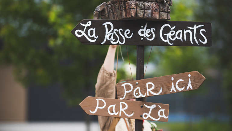 Un panneau directionnel artistique en bois affiche les inscriptions “La Passe des Géants”, “Par ici” et “Par là”. Au sommet du poteau se trouve une sculpture fantastique représentant une créature aux grands yeux avec une autre petite figure sur son dos. Une personne costumée est partiellement visible derrière le panneau, dans un décor extérieur verdoyant.