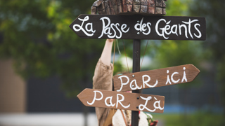 Un panneau directionnel artistique en bois affiche les inscriptions “La Passe des Géants”, “Par ici” et “Par là”. Au sommet du poteau se trouve une sculpture fantastique représentant une créature aux grands yeux avec une autre petite figure sur son dos. Une personne costumée est partiellement visible derrière le panneau, dans un décor extérieur verdoyant.
