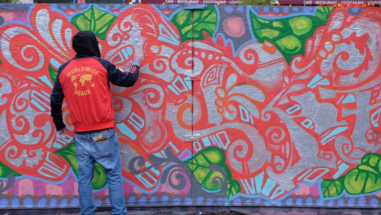 Personne peignant une murale colorée aux motifs abstraits et végétaux sur un mur extérieur.
