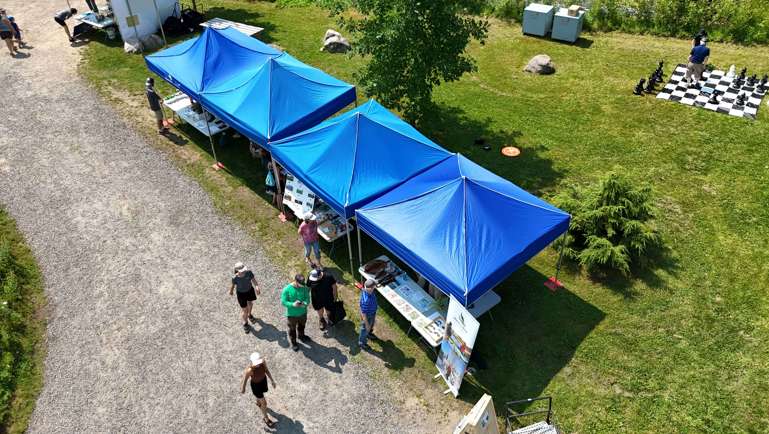 Vue aérienne d’un événement extérieur se déroulant dans un espace vert. Plusieurs kiosques sous des tentes bleues sont installés le long d’un chemin de gravier, où des visiteurs circulent et discutent. Des tables présentent du matériel d’information et des activités. À droite, une grande surface de jeu d’échecs géante est installée sur le gazon avec quelques personnes qui jouent. Journée ensoleillée avec arbres, rochers et ambiance conviviale de fête communautaire.