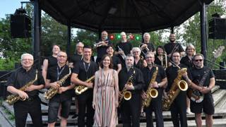Photo de groupe d’un ensemble de jazz posant devant une scène extérieure couverte. Les musiciens, vêtus majoritairement de noir, tiennent divers instruments comme des saxophones, trompettes et une basse électrique. Une femme au centre porte une robe rayée claire. Des arbres et des lumières de scène apparaissent en arrière-plan.