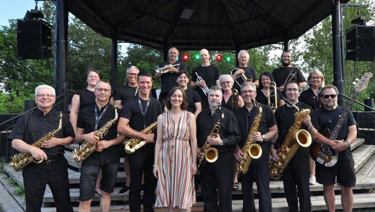 Photo de groupe d’un ensemble de jazz posant devant une scène extérieure couverte. Les musiciens, vêtus majoritairement de noir, tiennent divers instruments comme des saxophones, trompettes et une basse électrique. Une femme au centre porte une robe rayée claire. Des arbres et des lumières de scène apparaissent en arrière-plan.