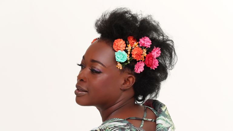 Portrait d’une femme portant une robe à motifs tropicaux verts et roses avec des volants. Elle pose de profil devant un fond clair, les cheveux relevés et ornés de fleurs colorées. Son expression est douce et élégante, dans une ambiance lumineuse et artistique.