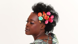 Portrait d’une femme portant une robe à motifs tropicaux verts et roses avec des volants. Elle pose de profil devant un fond clair, les cheveux relevés et ornés de fleurs colorées. Son expression est douce et élégante, dans une ambiance lumineuse et artistique.
