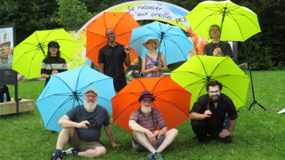 Groupe de sept personnes posant dans un parc avec des parapluies colorés vert lime, orange et bleu. Certaines personnes sont debout et d’autres assises sur l’herbe. En arrière-plan, une installation artistique colorée et des arbres entourent la scène.