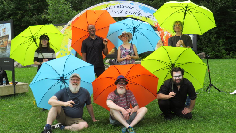Groupe de sept personnes posant dans un parc avec des parapluies colorés vert lime, orange et bleu. Certaines personnes sont debout et d’autres assises sur l’herbe. En arrière-plan, une installation artistique colorée et des arbres entourent la scène.