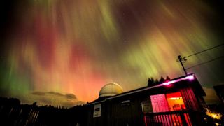 Ciel nocturne illuminé par des aurores boréales vertes, rouges et jaunes au-dessus d’un petit observatoire. Le bâtiment sombre possède une coupole blanche et une porte éclairée en rouge, tandis que des arbres et des collines se dessinent à l’horizon.